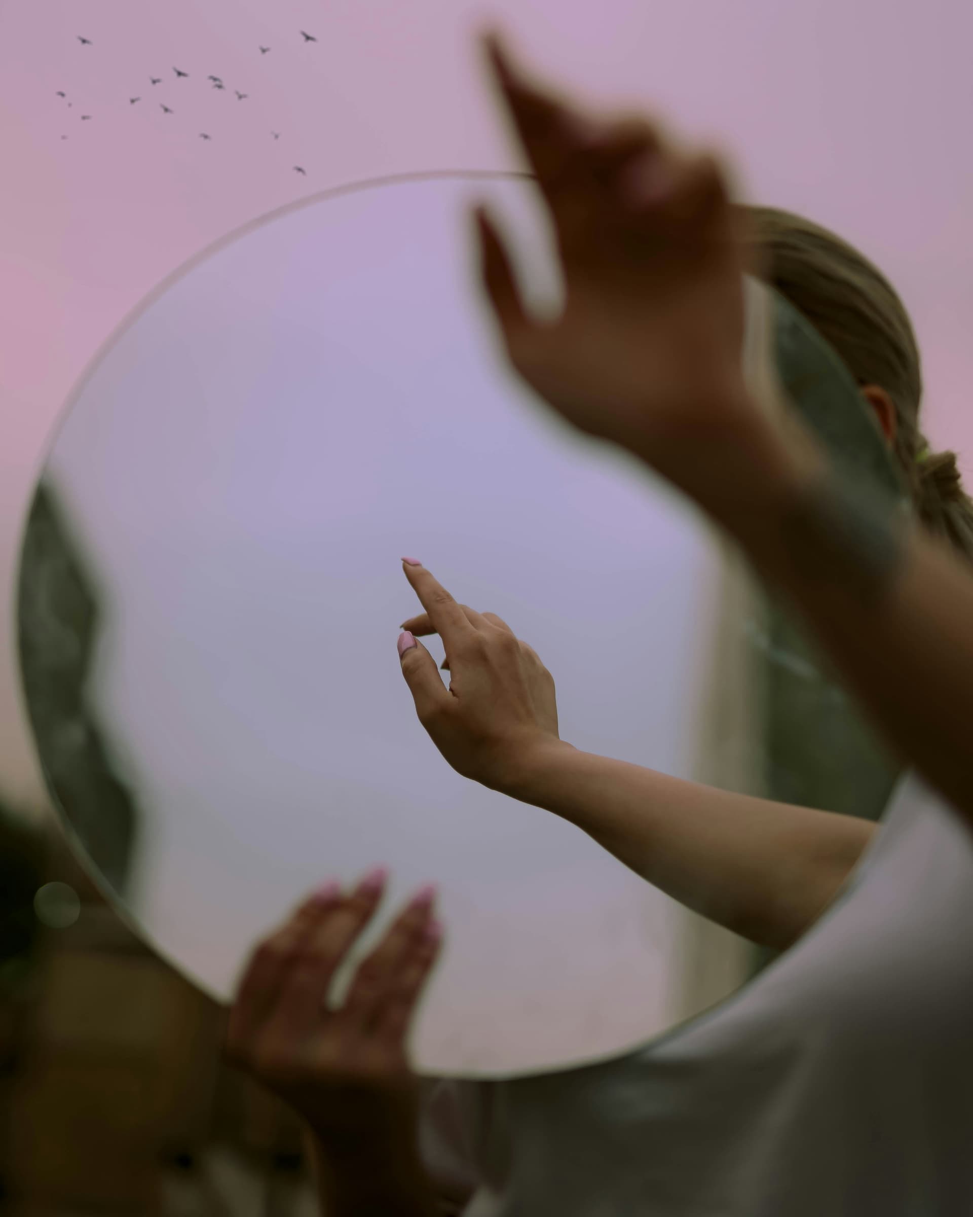 En hand som rör vid en rund spegel mot en rosa himmel